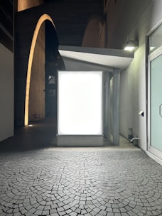 An illuminated billboard stand is placed on a cobblestone pavement in a dimly lit, narrow alley. The scene is bordered by high walls and an overhanging archway, with the glow from the billboard casting light onto the surrounding environment.