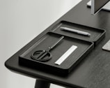 A desktop setup featuring a sleek, minimalistic design with a focus on organization. Two black trays hold a pair of scissors, a pen, and a small ruler. The table and trays have a modern, dark finish, and a portion of a monitor is visible in the corner.