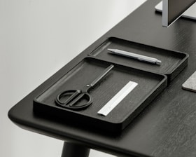 A desktop setup featuring a sleek, minimalistic design with a focus on organization. Two black trays hold a pair of scissors, a pen, and a small ruler. The table and trays have a modern, dark finish, and a portion of a monitor is visible in the corner.