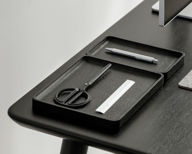 A desktop setup featuring a sleek, minimalistic design with a focus on organization. Two black trays hold a pair of scissors, a pen, and a small ruler. The table and trays have a modern, dark finish, and a portion of a monitor is visible in the corner.