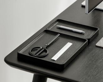 A desktop setup featuring a sleek, minimalistic design with a focus on organization. Two black trays hold a pair of scissors, a pen, and a small ruler. The table and trays have a modern, dark finish, and a portion of a monitor is visible in the corner.