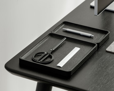 A desktop setup featuring a sleek, minimalistic design with a focus on organization. Two black trays hold a pair of scissors, a pen, and a small ruler. The table and trays have a modern, dark finish, and a portion of a monitor is visible in the corner.