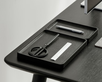 A desktop setup featuring a sleek, minimalistic design with a focus on organization. Two black trays hold a pair of scissors, a pen, and a small ruler. The table and trays have a modern, dark finish, and a portion of a monitor is visible in the corner.