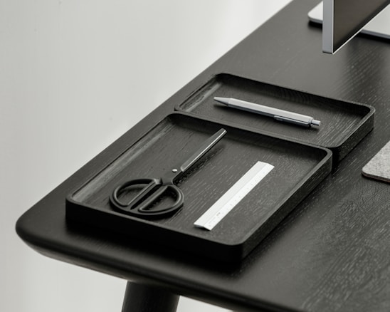A desktop setup featuring a sleek, minimalistic design with a focus on organization. Two black trays hold a pair of scissors, a pen, and a small ruler. The table and trays have a modern, dark finish, and a portion of a monitor is visible in the corner.
