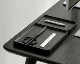 A desktop setup featuring a sleek, minimalistic design with a focus on organization. Two black trays hold a pair of scissors, a pen, and a small ruler. The table and trays have a modern, dark finish, and a portion of a monitor is visible in the corner.