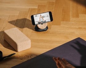 A smartphone is resting on a wooden stand, displaying a video of a person doing yoga. The setup is on a wooden floor with a yoga block and a black yoga mat nearby. A hand is visible, suggesting someone is preparing to follow the workout.