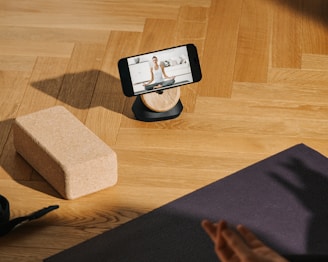 A smartphone is resting on a wooden stand, displaying a video of a person doing yoga. The setup is on a wooden floor with a yoga block and a black yoga mat nearby. A hand is visible, suggesting someone is preparing to follow the workout.