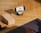 A smartphone is resting on a wooden stand, displaying a video of a person doing yoga. The setup is on a wooden floor with a yoga block and a black yoga mat nearby. A hand is visible, suggesting someone is preparing to follow the workout.