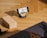 A smartphone is resting on a wooden stand, displaying a video of a person doing yoga. The setup is on a wooden floor with a yoga block and a black yoga mat nearby. A hand is visible, suggesting someone is preparing to follow the workout.
