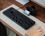 A computer desk setup featuring an editing console with multiple dials and buttons, accompanied by a digital camera and a wireless mouse. The console is placed on a felt mat on a wooden desk surface, with a part of a person's hand interacting with the mouse.