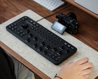 A computer desk setup featuring an editing console with multiple dials and buttons, accompanied by a digital camera and a wireless mouse. The console is placed on a felt mat on a wooden desk surface, with a part of a person's hand interacting with the mouse.