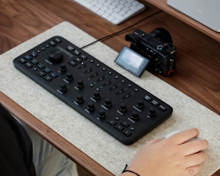A computer desk setup featuring an editing console with multiple dials and buttons, accompanied by a digital camera and a wireless mouse. The console is placed on a felt mat on a wooden desk surface, with a part of a person's hand interacting with the mouse.