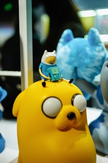 A small figurine sitting atop a larger yellow character with a distinctive round face and large eyes. The smaller figure resembles a humanoid character wearing a white hat with pointed ears and holding a small device with a face. The background includes a blurred blue figure.