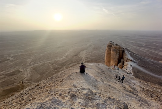 A person sits at the edge of a rocky cliff overlooking a vast, barren desert stretching into the horizon under a hazy sky with the sun near setting. Nearby, other people explore the rugged terrain and smaller rocky formations.