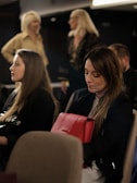 A group of individuals is seated, with one woman prominently holding a red handbag. The setting appears to be a formal indoor environment with soft lighting. Two other women are conversing in the background, standing with blurred features.
