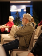 An older man with a beard and glasses sits attentively in a conference room, surrounded by empty chairs. He is wearing a green jacket and blue jeans. In the background, a woman with white hair in a red shirt appears to be seated. The room has a modern ambiance with overhead lighting and some decorative plants.