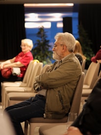 An older man with a beard and glasses sits attentively in a conference room, surrounded by empty chairs. He is wearing a green jacket and blue jeans. In the background, a woman with white hair in a red shirt appears to be seated. The room has a modern ambiance with overhead lighting and some decorative plants.