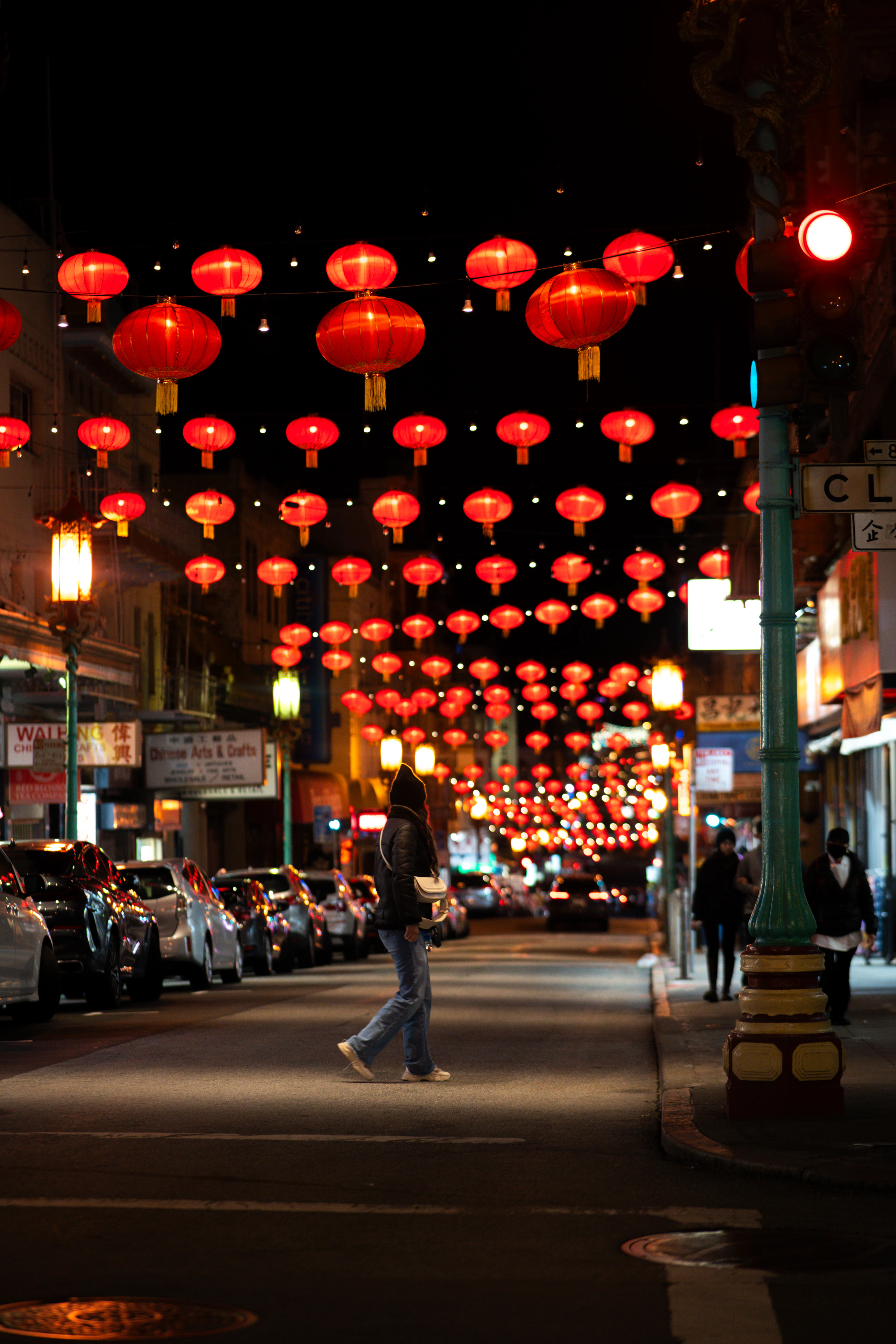 Rows of red paper lanterns strung above a San Francisco Chinatown street at night, a lone pedestrian crossing beneath them