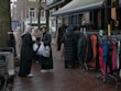 A group of women dressed in various coats, standing near a street market stall with racks of colorful clothing. Some of the women are carrying shopping bags. The pavement is wet, suggesting recent rainfall, and mannequins are visible, showcasing some of the clothing.