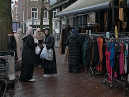 A group of women dressed in various coats, standing near a street market stall with racks of colorful clothing. Some of the women are carrying shopping bags. The pavement is wet, suggesting recent rainfall, and mannequins are visible, showcasing some of the clothing.