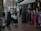 A group of women dressed in various coats, standing near a street market stall with racks of colorful clothing. Some of the women are carrying shopping bags. The pavement is wet, suggesting recent rainfall, and mannequins are visible, showcasing some of the clothing.
