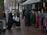 A group of women dressed in various coats, standing near a street market stall with racks of colorful clothing. Some of the women are carrying shopping bags. The pavement is wet, suggesting recent rainfall, and mannequins are visible, showcasing some of the clothing.