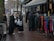 A group of women dressed in various coats, standing near a street market stall with racks of colorful clothing. Some of the women are carrying shopping bags. The pavement is wet, suggesting recent rainfall, and mannequins are visible, showcasing some of the clothing.