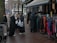 A group of women dressed in various coats, standing near a street market stall with racks of colorful clothing. Some of the women are carrying shopping bags. The pavement is wet, suggesting recent rainfall, and mannequins are visible, showcasing some of the clothing.