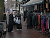A group of women dressed in various coats, standing near a street market stall with racks of colorful clothing. Some of the women are carrying shopping bags. The pavement is wet, suggesting recent rainfall, and mannequins are visible, showcasing some of the clothing.