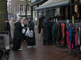 A group of women dressed in various coats, standing near a street market stall with racks of colorful clothing. Some of the women are carrying shopping bags. The pavement is wet, suggesting recent rainfall, and mannequins are visible, showcasing some of the clothing.