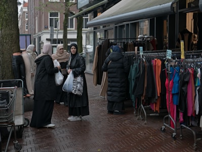 A group of women dressed in various coats, standing near a street market stall with racks of colorful clothing. Some of the women are carrying shopping bags. The pavement is wet, suggesting recent rainfall, and mannequins are visible, showcasing some of the clothing.