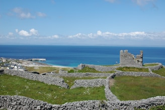 A scenic landscape featuring a historical stone structure amid grassy fields and numerous stone walls, set against a backdrop of vibrant blue ocean and a partly cloudy sky. A small village with houses is visible near the coast alongside a pier extending into the water.