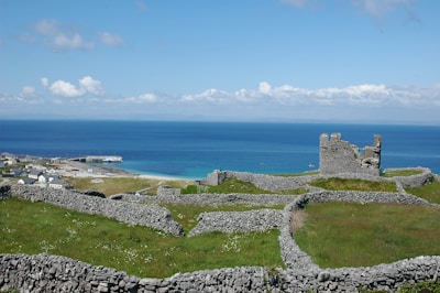 A scenic landscape featuring a historical stone structure amid grassy fields and numerous stone walls, set against a backdrop of vibrant blue ocean and a partly cloudy sky. A small village with houses is visible near the coast alongside a pier extending into the water.