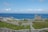 A scenic landscape featuring a historical stone structure amid grassy fields and numerous stone walls, set against a backdrop of vibrant blue ocean and a partly cloudy sky. A small village with houses is visible near the coast alongside a pier extending into the water.