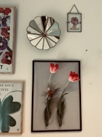 A collection of framed art pieces hung on a wall, including a decorative mirror with a scalloped edge. The main framed piece features two vibrant red tulips with a small toy mouse nestled between the stems. Other frames contain floral and abstract designs, one with text in French.