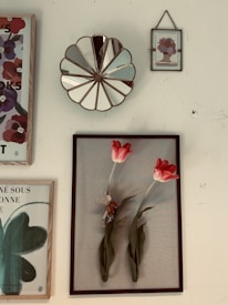 A collection of framed art pieces hung on a wall, including a decorative mirror with a scalloped edge. The main framed piece features two vibrant red tulips with a small toy mouse nestled between the stems. Other frames contain floral and abstract designs, one with text in French.
