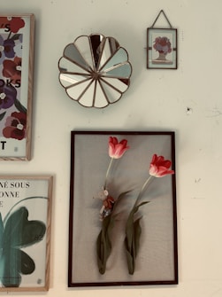 A collection of framed art pieces hung on a wall, including a decorative mirror with a scalloped edge. The main framed piece features two vibrant red tulips with a small toy mouse nestled between the stems. Other frames contain floral and abstract designs, one with text in French.