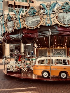 A vintage carousel in an urban setting features ornately decorated panels with angelic figures and old-style photographs. It includes carousel horses and a retro-style vehicle ride, with intricate designs in aqua and cream colors. The surrounding architecture shows brick buildings and shops.