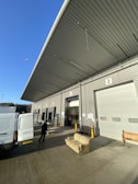 A large warehouse loading area with numbered doors, open to the outside. A van is parked nearby with its back doors open, and a person is transporting a dolly or cart. A striped sofa is placed in front of the loading dock, with a wooden bench visible nearby. The sky is clear and blue, indicating a sunny day. The long, overhanging roof is angled steeply and partially casts a shadow over the area.