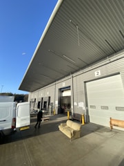 A large warehouse loading area with numbered doors, open to the outside. A van is parked nearby with its back doors open, and a person is transporting a dolly or cart. A striped sofa is placed in front of the loading dock, with a wooden bench visible nearby. The sky is clear and blue, indicating a sunny day. The long, overhanging roof is angled steeply and partially casts a shadow over the area.