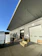 A large warehouse loading area with numbered doors, open to the outside. A van is parked nearby with its back doors open, and a person is transporting a dolly or cart. A striped sofa is placed in front of the loading dock, with a wooden bench visible nearby. The sky is clear and blue, indicating a sunny day. The long, overhanging roof is angled steeply and partially casts a shadow over the area.