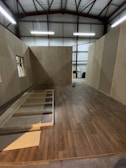 A partially constructed room inside a warehouse, featuring wooden floors and walls with exposed beams on the ceiling. Unfinished wooden frames are visible on the floor, with some construction tools in the background.