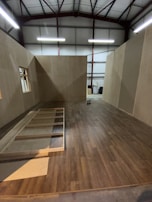 A partially constructed room inside a warehouse, featuring wooden floors and walls with exposed beams on the ceiling. Unfinished wooden frames are visible on the floor, with some construction tools in the background.