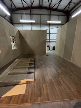 A partially constructed room inside a warehouse, featuring wooden floors and walls with exposed beams on the ceiling. Unfinished wooden frames are visible on the floor, with some construction tools in the background.