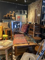 A cozy interior filled with various vintage furniture and decor items, including a woven tapestry, wooden tables and chairs, and decorative items like wooden horse sculptures and retro signs. The space has an eclectic and rustic charm, with colorful patterns and textures throughout.