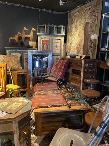 A cozy interior filled with various vintage furniture and decor items, including a woven tapestry, wooden tables and chairs, and decorative items like wooden horse sculptures and retro signs. The space has an eclectic and rustic charm, with colorful patterns and textures throughout.
