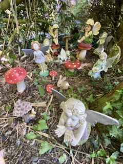 A collection of decorative fairy statues is arranged on the forest floor, surrounded by foliage. The fairies have delicate wings and sit on or near oversized red and white toadstools. There are also other small statues, including a rabbit. The area is shaded by trees, creating a whimsical and magical atmosphere.