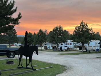 A silhouette of a cowboy on horseback stands prominently against a backdrop of an RV park. The sky displays vibrant hues of orange and pink as the sun sets, casting a warm glow over the landscape. Several recreational vehicles and trees are arranged along a gravel path. The atmosphere is calm and serene, with a mix of natural and manmade elements.