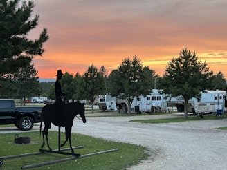 A silhouette of a cowboy on horseback stands prominently against a backdrop of an RV park. The sky displays vibrant hues of orange and pink as the sun sets, casting a warm glow over the landscape. Several recreational vehicles and trees are arranged along a gravel path. The atmosphere is calm and serene, with a mix of natural and manmade elements.