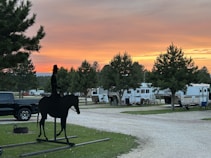 A silhouette of a cowboy on horseback stands prominently against a backdrop of an RV park. The sky displays vibrant hues of orange and pink as the sun sets, casting a warm glow over the landscape. Several recreational vehicles and trees are arranged along a gravel path. The atmosphere is calm and serene, with a mix of natural and manmade elements.